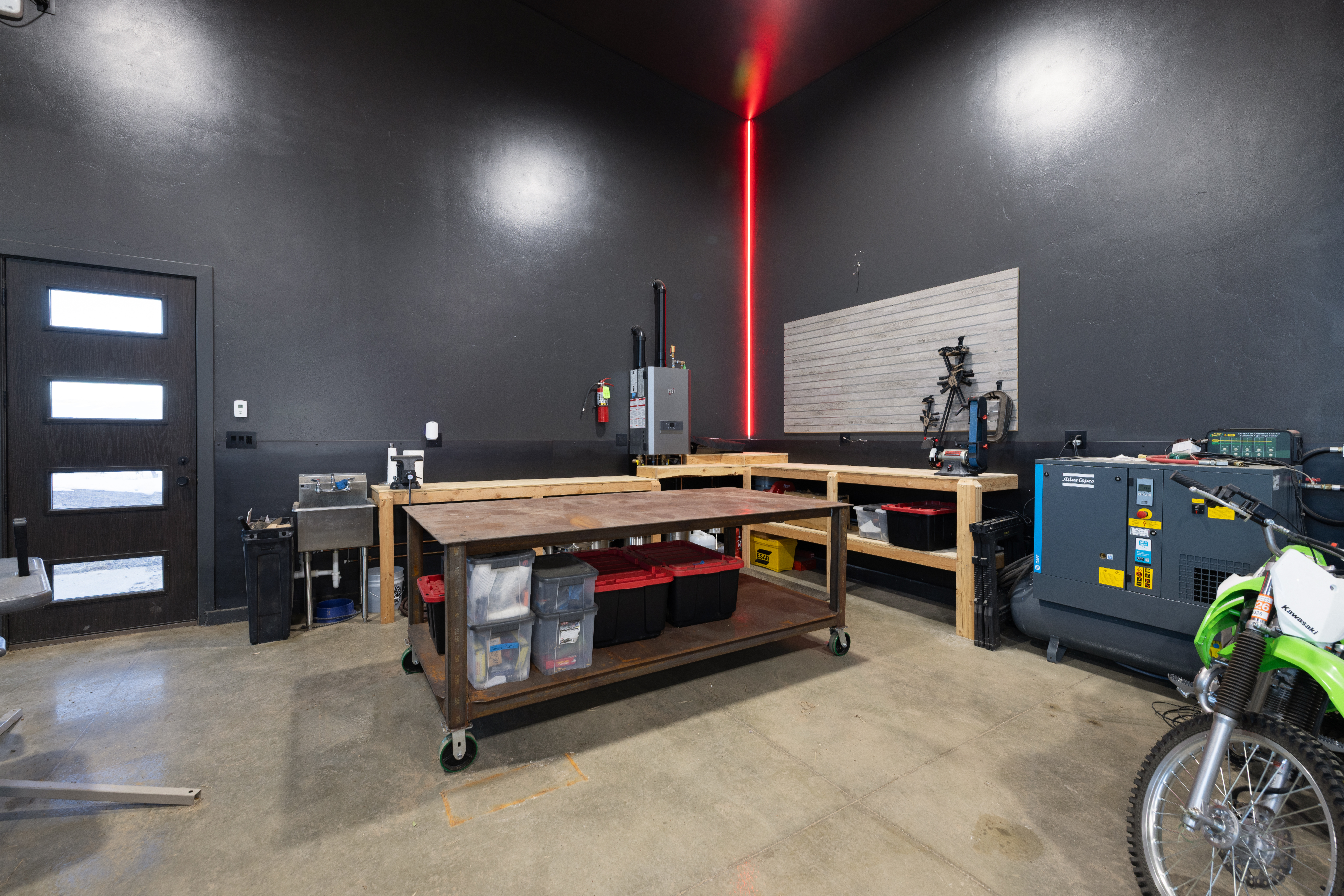 This interior garage workspace is well-equipped for projects, featuring a large, sturdy mobile metal workbench and extensive wooden shelving. A sleek, black-walled aesthetic is highlighted by a modern vertical red LED accent light in the corner. The setup includes a utility sink, clear storage bins, and heavy-duty machinery, making it an ideal functional space for DIY enthusiasts or professional use.