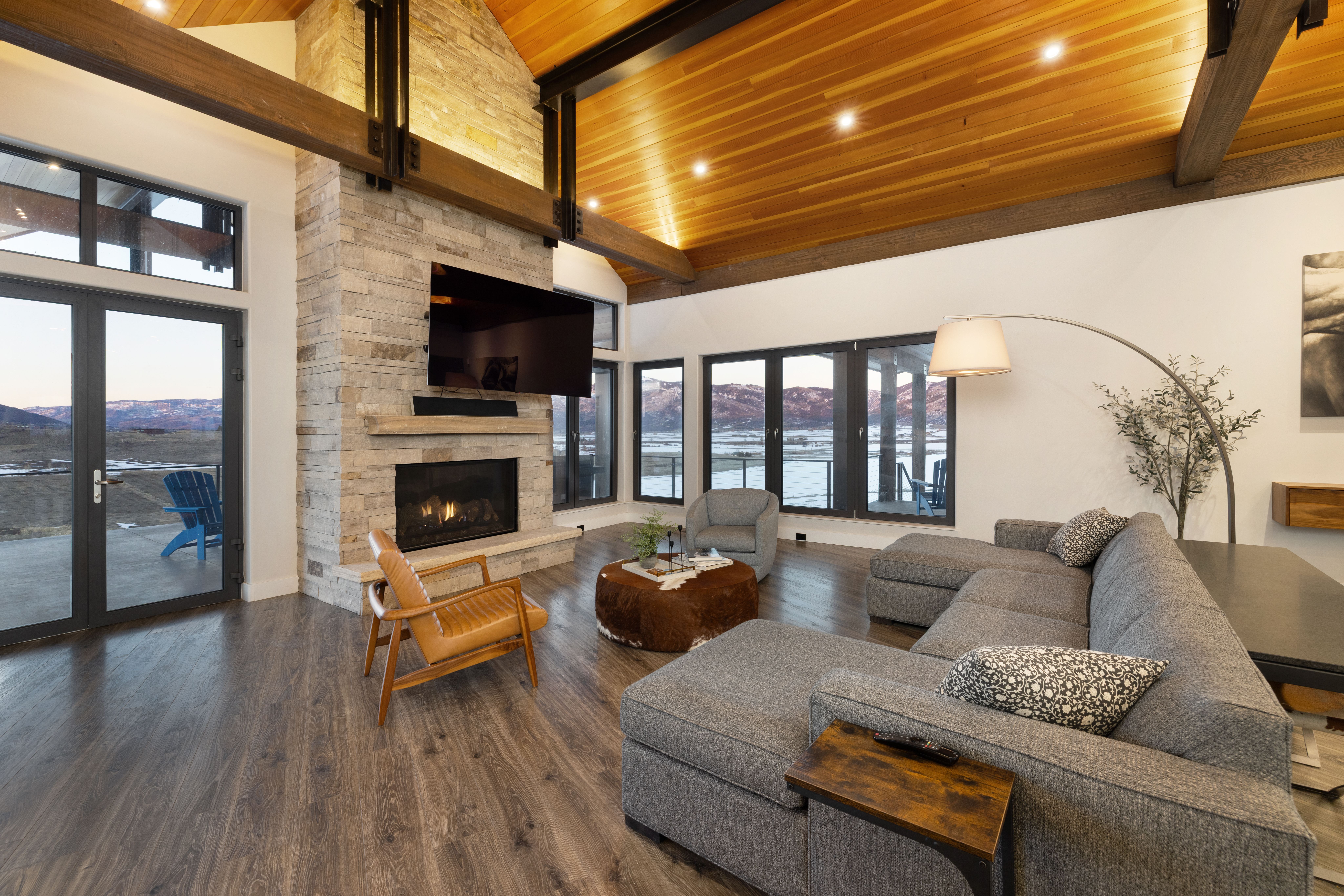 This stunning contemporary living room features a vaulted wood-paneled ceiling, exposed structural beams, and a floor-to-ceiling flagstone fireplace. The space is comfortably furnished with a plush grey sectional, a circular cowhide ottoman, and a mid-century inspired leather chair. Large windows and glass doors frame breathtaking mountain views, seamlessly blending modern luxury with a cozy, rustic aesthetic.