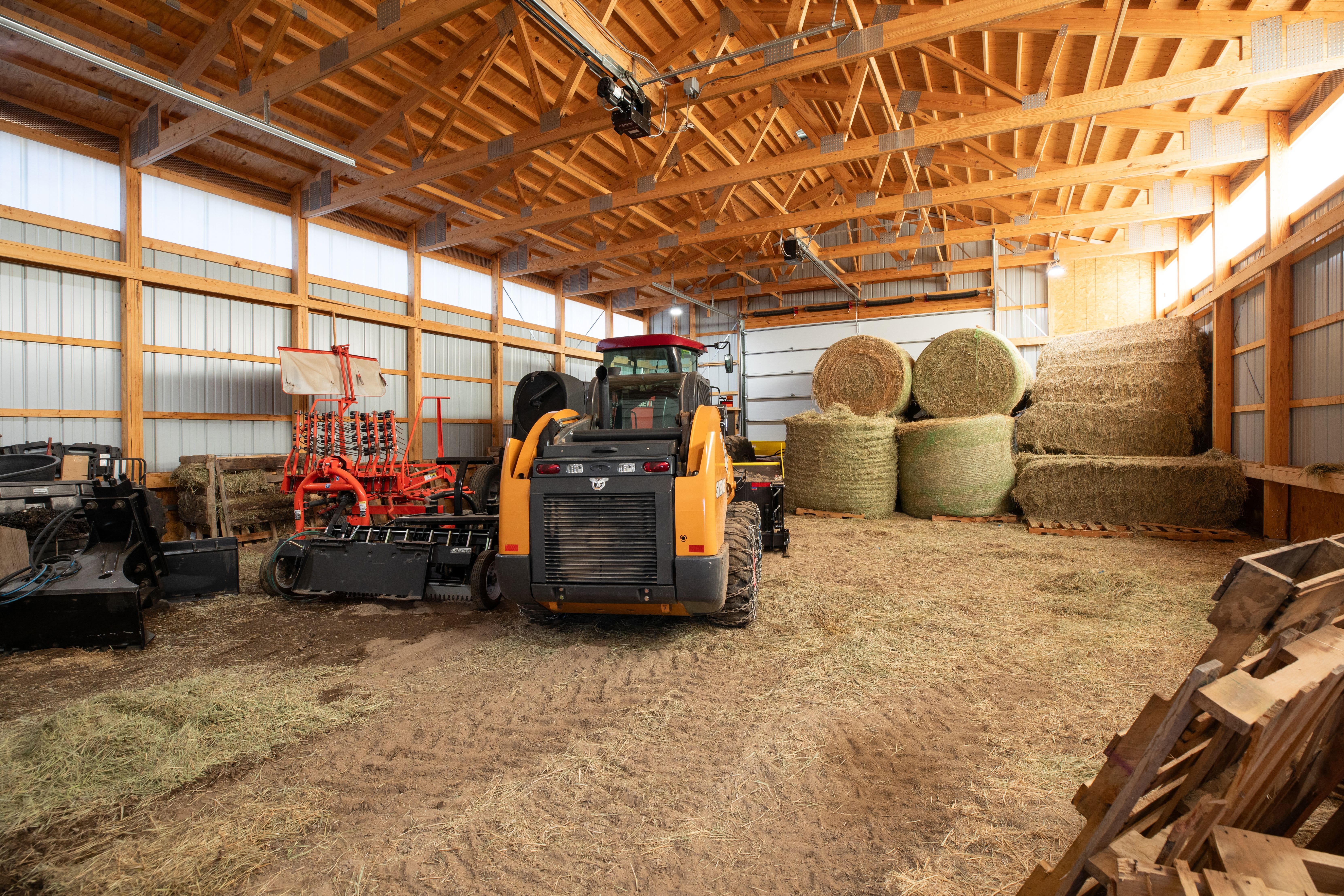 This spacious interior agricultural outbuilding, such as a pole barn, features exposed wooden trusses and metal siding, providing a functional setting for equipment storage. The space is used for housing farm machinery, including a skid steer and implements, as well as storing stacked round hay bales. It offers a practical and versatile workspace suited for farming and ranching operations.