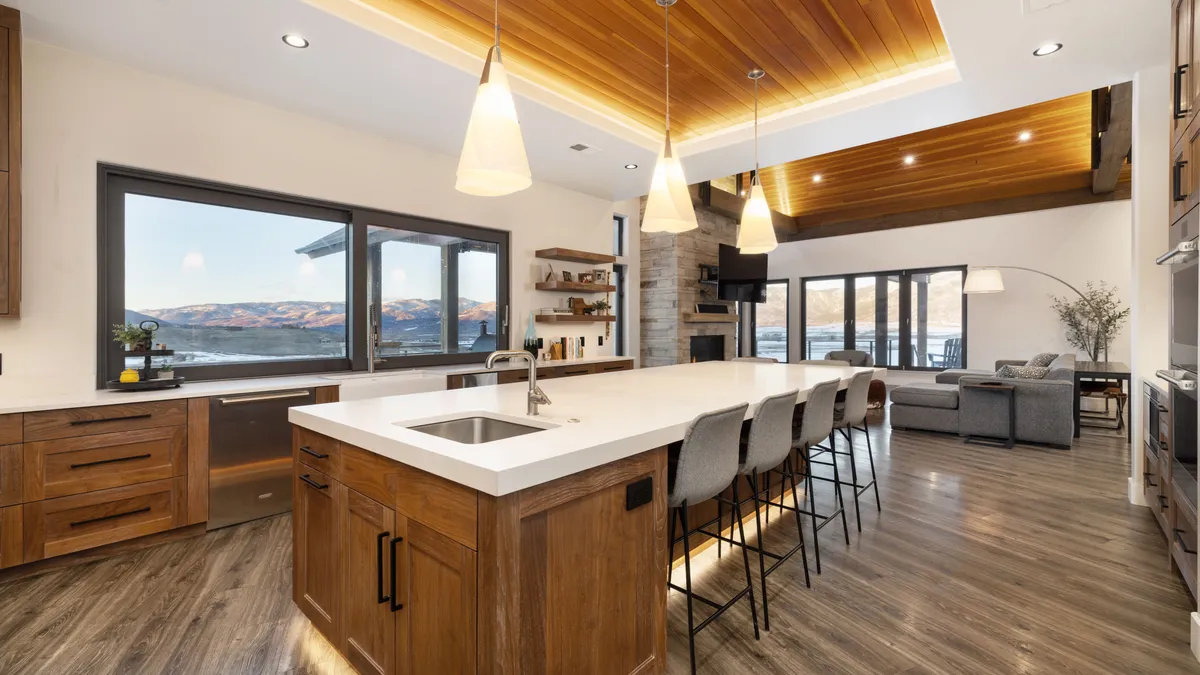 This modern open-concept kitchen features warm wood cabinetry contrasted by sleek white quartz countertops and a large central island with bar seating. The space flows seamlessly into a living area, accented by a stunning cedar plank tray ceiling and oversized windows with panoramic mountain views. The design combines rustic warmth with contemporary luxury, highlighted by stylish pendant lighting and stainless steel appliances.