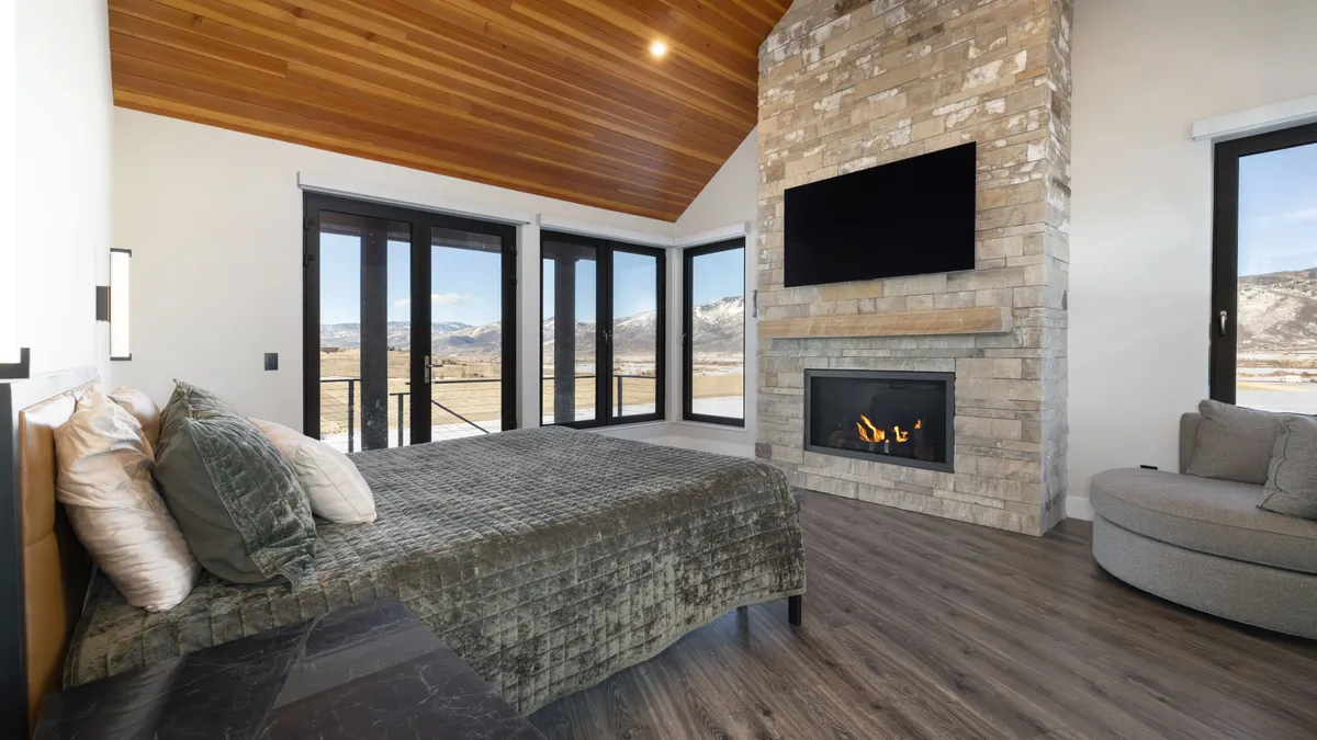 This spacious primary bedroom features a modern, rustic aesthetic with a dramatic vaulted wood-paneled ceiling and a large stone fireplace as a centerpiece. The room includes a cozy bed, plush circular seating, and expansive floor-to-ceiling windows that provide panoramic mountain views, creating a luxurious and serene atmosphere.