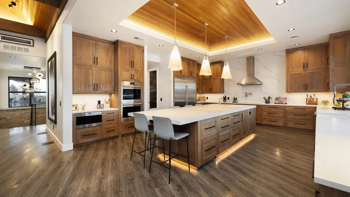 This stunning contemporary kitchen features rich, warm wood cabinetry paired with crisp white quartz countertops and a large central island with bar seating. The space is highlighted by recessed lighting and elegant pendant fixtures, set against a beautifully vaulted wood-paneled ceiling. Modern stainless steel appliances and clean, minimalist lines create a sophisticated and cohesive open-concept cooking and entertaining area.