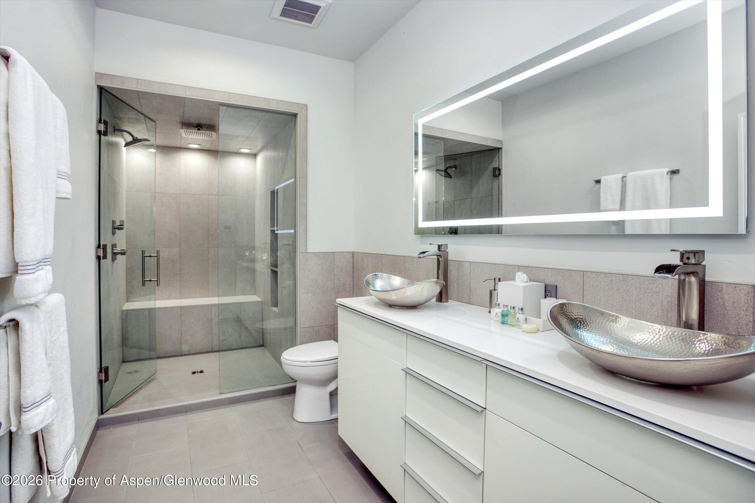 This modern primary bathroom features a sleek double vanity with metallic vessel sinks and a large, illuminated rectangular mirror. To the left, a spacious walk-in shower with glass walls and a built-in bench is finished in neutral-toned tile. The room is bright and contemporary, utilizing clean lines and a minimalist aesthetic to create a spa-like atmosphere.