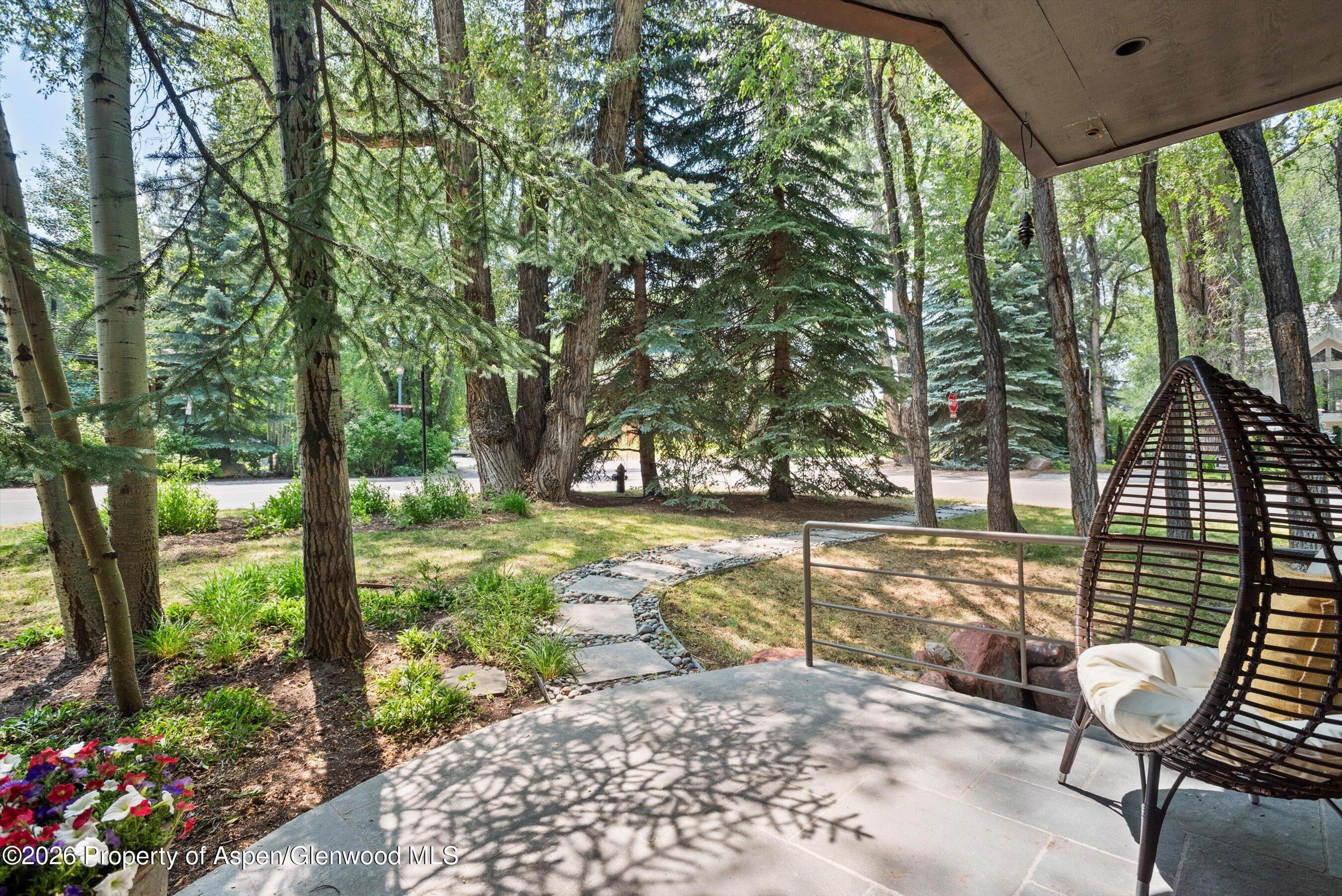 This image captures a serene, covered patio space featuring a modern hanging egg chair and a stone-tiled floor. The perspective looks out from the shaded porch toward a lush, wooded yard with a winding stone pathway leading through the grass. The scene is framed by mature evergreen and deciduous trees, creating a peaceful and private outdoor retreat.