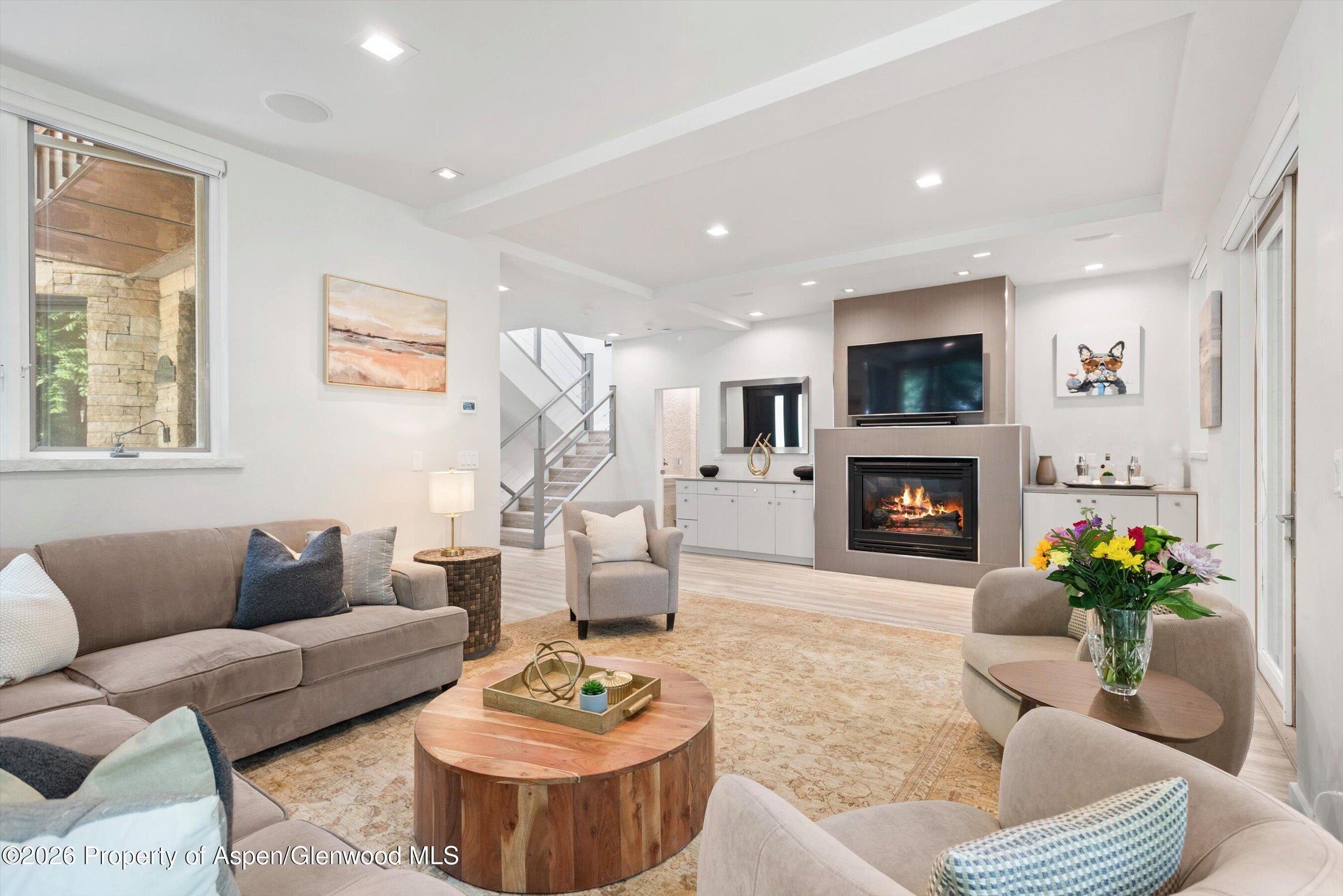 This open-concept living area features light-toned wood flooring and a contemporary aesthetic with a neutral color palette. A prominent fireplace with a tall, sleek surround serves as the focal point, topped by a mounted television, while a comfortable armchair sits on a textured area rug in the foreground. In the background, a staircase with metal railings leads to an upper level, and a glimpse of a bright, modern kitchen is visible through the open floor plan.