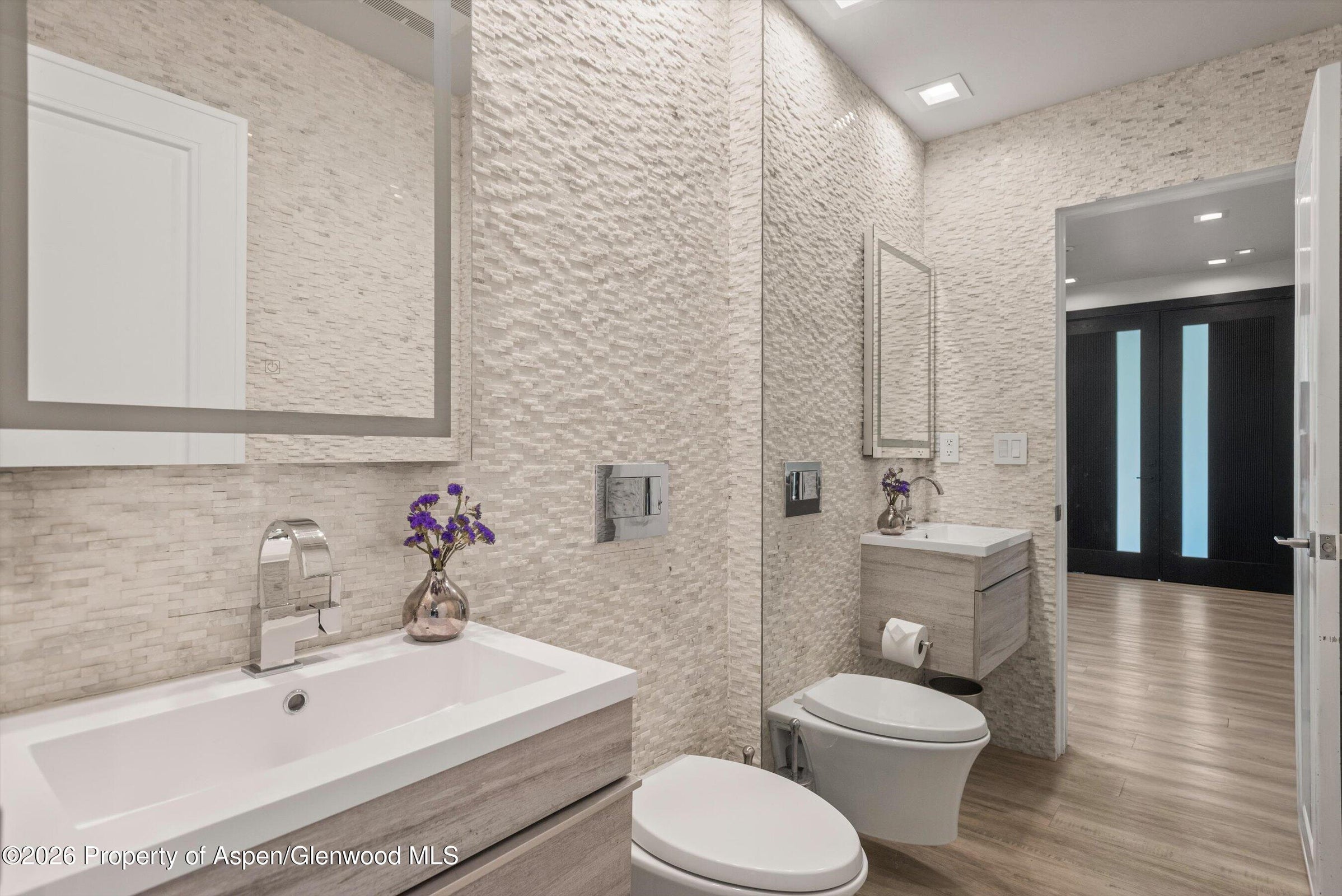 This modern powder room features a textured stone wall that adds depth and visual interest to the space. It includes two floating vanities with light wood finishes, wall-mounted toilets, and sleek chrome fixtures. The room is illuminated by recessed lighting and mirrors, creating a clean, contemporary aesthetic that feels both compact and sophisticated.