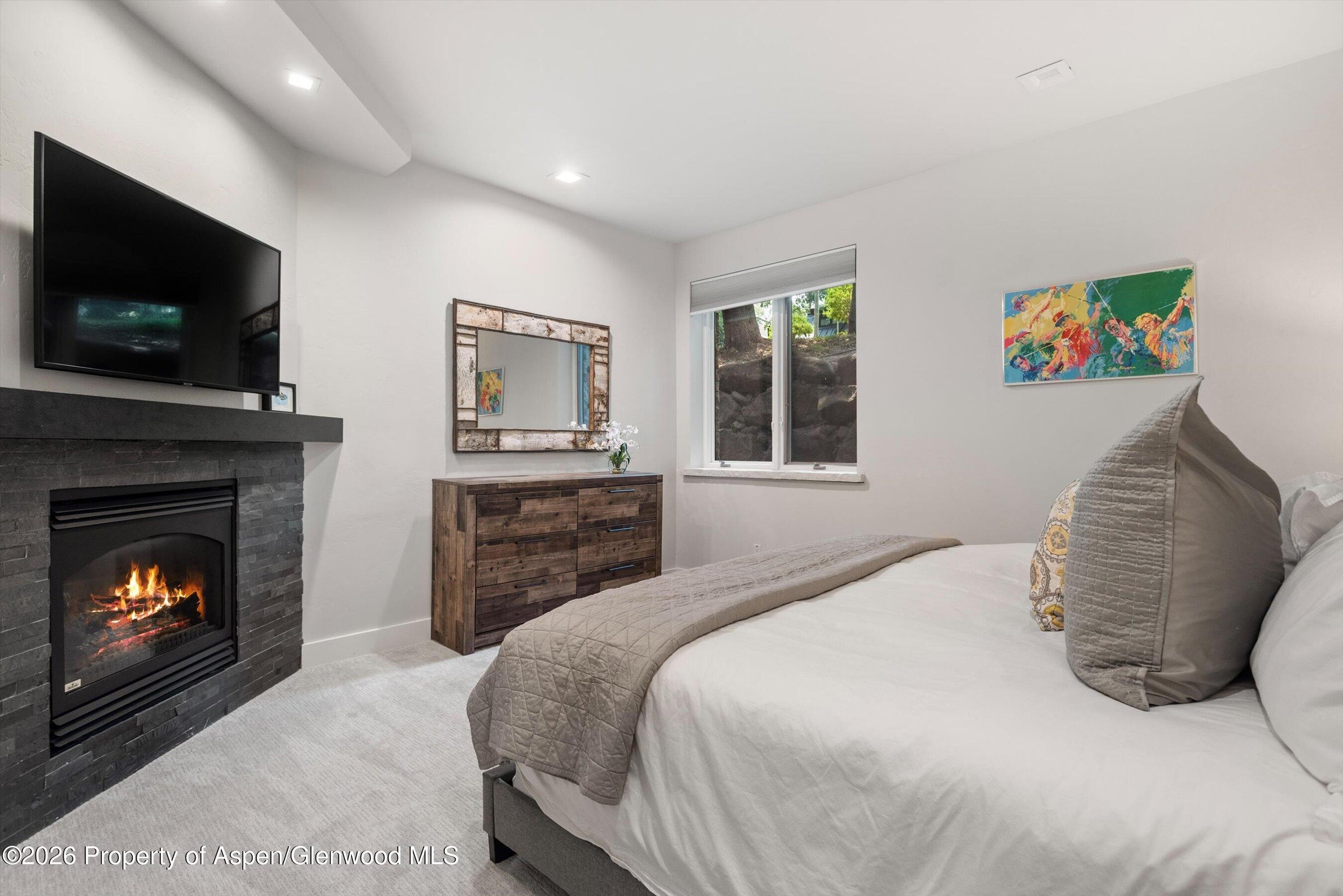 This spacious primary bedroom features a vaulted ceiling with recessed lighting and a neutral color palette that enhances the airy atmosphere. The room includes a cozy seating area with a sofa and a live-edge wood coffee table, alongside a private balcony accessible through a glass door. Large windows provide abundant natural light and scenic views of the surrounding trees, creating a serene and tranquil retreat.