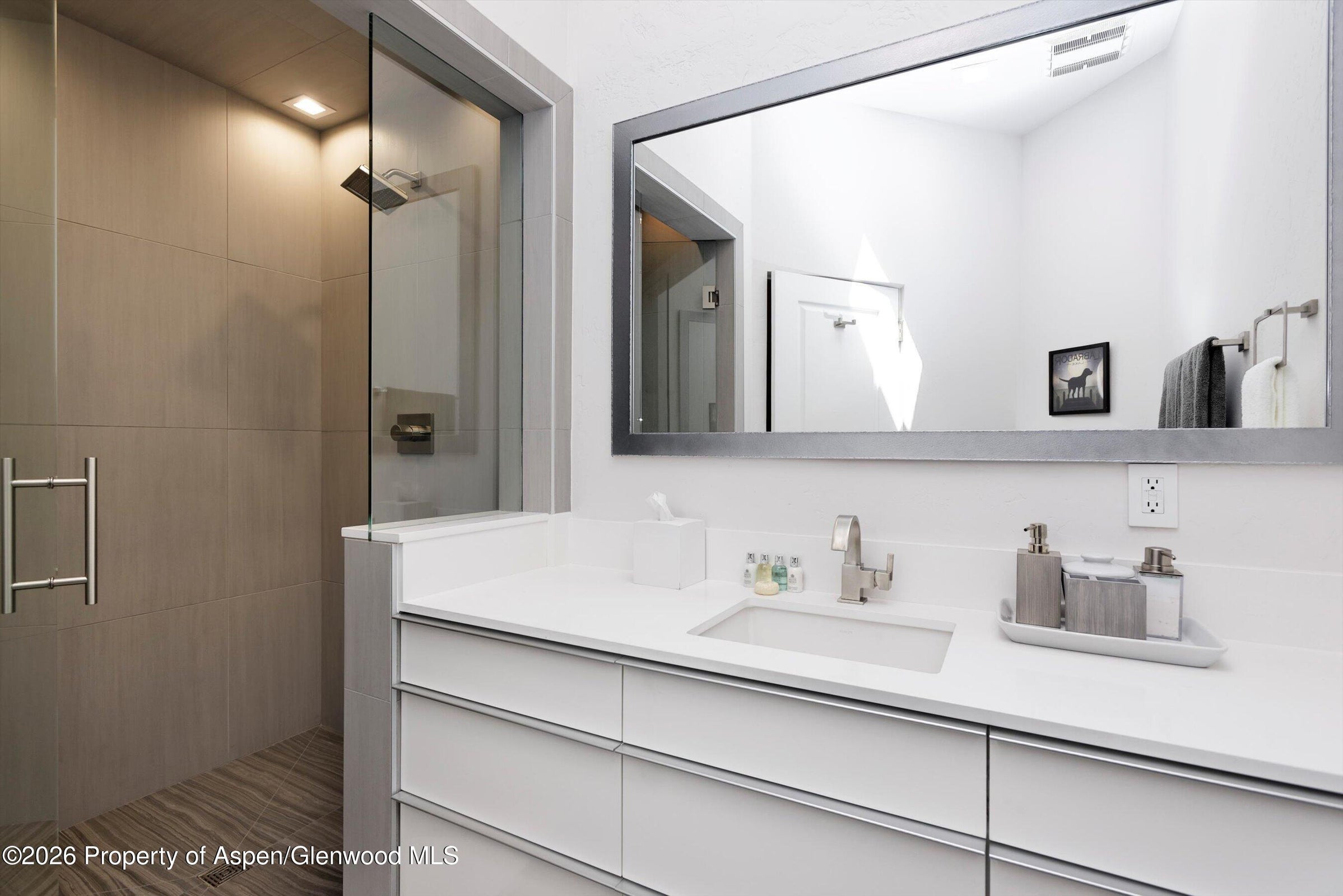 This modern bathroom features a sleek vanity with a white countertop, a rectangular undermount sink, and minimalist cabinetry. To the left, a glass-enclosed walk-in shower showcases neutral-toned wall tiles and a rainfall showerhead. A large, rectangular mirror with a dark frame hangs above the vanity, reflecting the bright, clean aesthetic of the space.