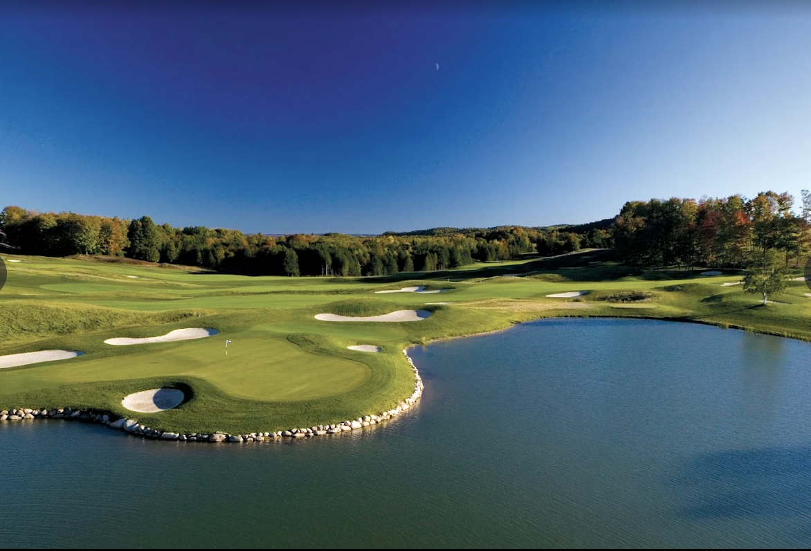 This aerial view showcases a beautifully landscaped golf course with lush green fairways, strategically placed sand traps, and a serene water feature. The course is surrounded by mature trees, creating a picturesque and tranquil setting. The clear blue sky enhances the overall appeal, suggesting a perfect day for golfing.