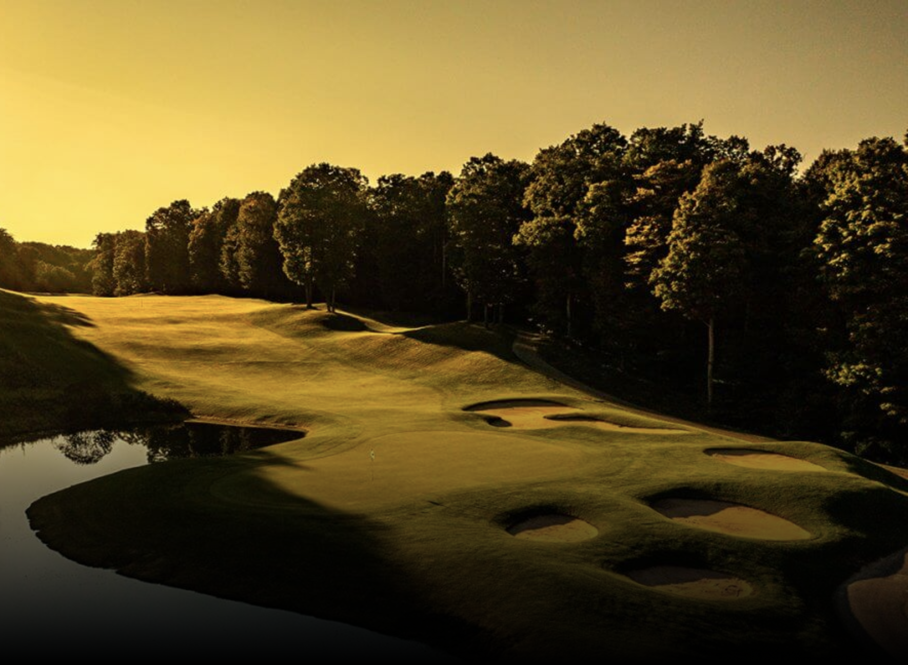 This image showcases a beautifully landscaped golf course with lush green fairways, strategically placed sand traps, and a serene pond. The course is framed by mature trees, creating a sense of privacy and tranquility. The golden hour lighting enhances the natural beauty, making it an appealing feature for potential buyers seeking a luxurious and recreational property.