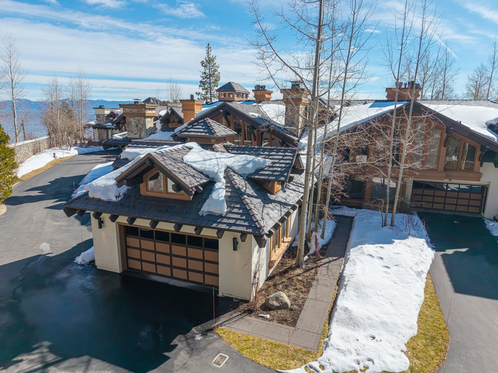 This aerial view showcases a luxurious mountain home with a multi-car garage, a dark asphalt driveway, and a roof partially covered in snow. The architecture features a blend of natural materials and modern design, with multiple gabled roofs and stone accents. The surrounding landscape includes bare trees and patches of snow, suggesting a serene, upscale setting.