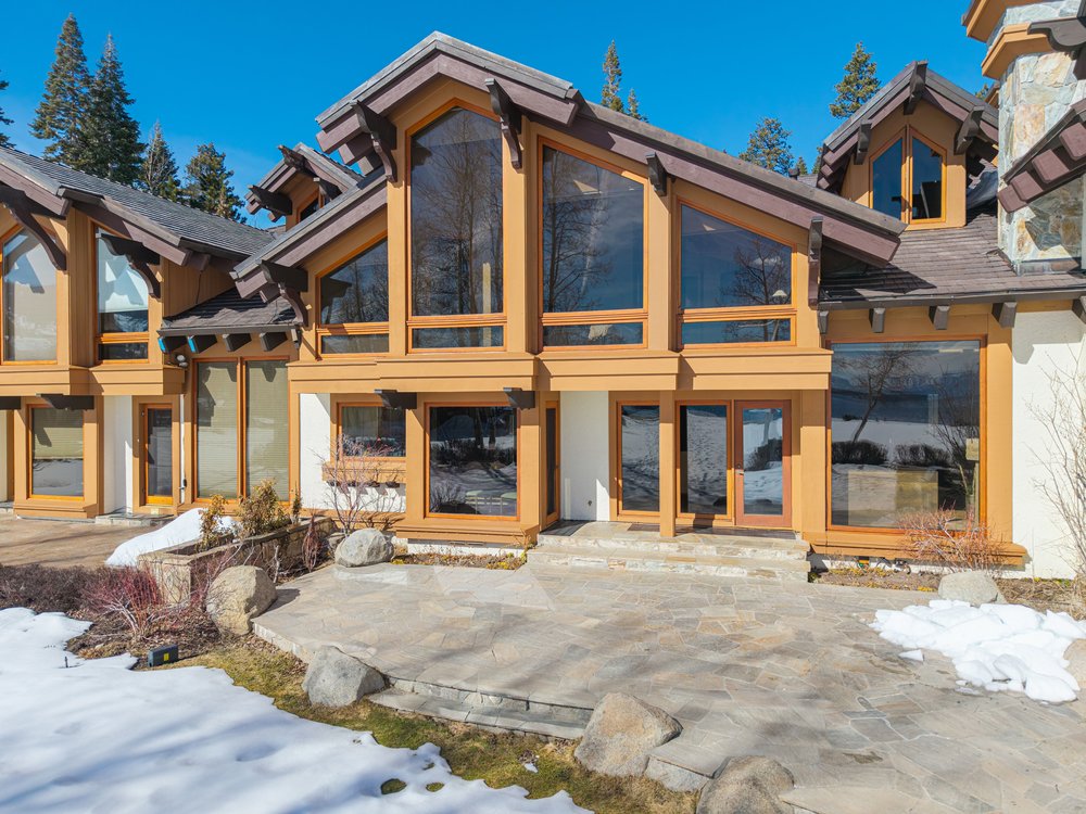 This is a front view of a luxurious multi-story home with large windows and a stone patio. The exterior features a combination of wood and light-colored siding, with a roof that has multiple gables. Patches of snow are visible on the ground, suggesting a mountain or ski resort location.