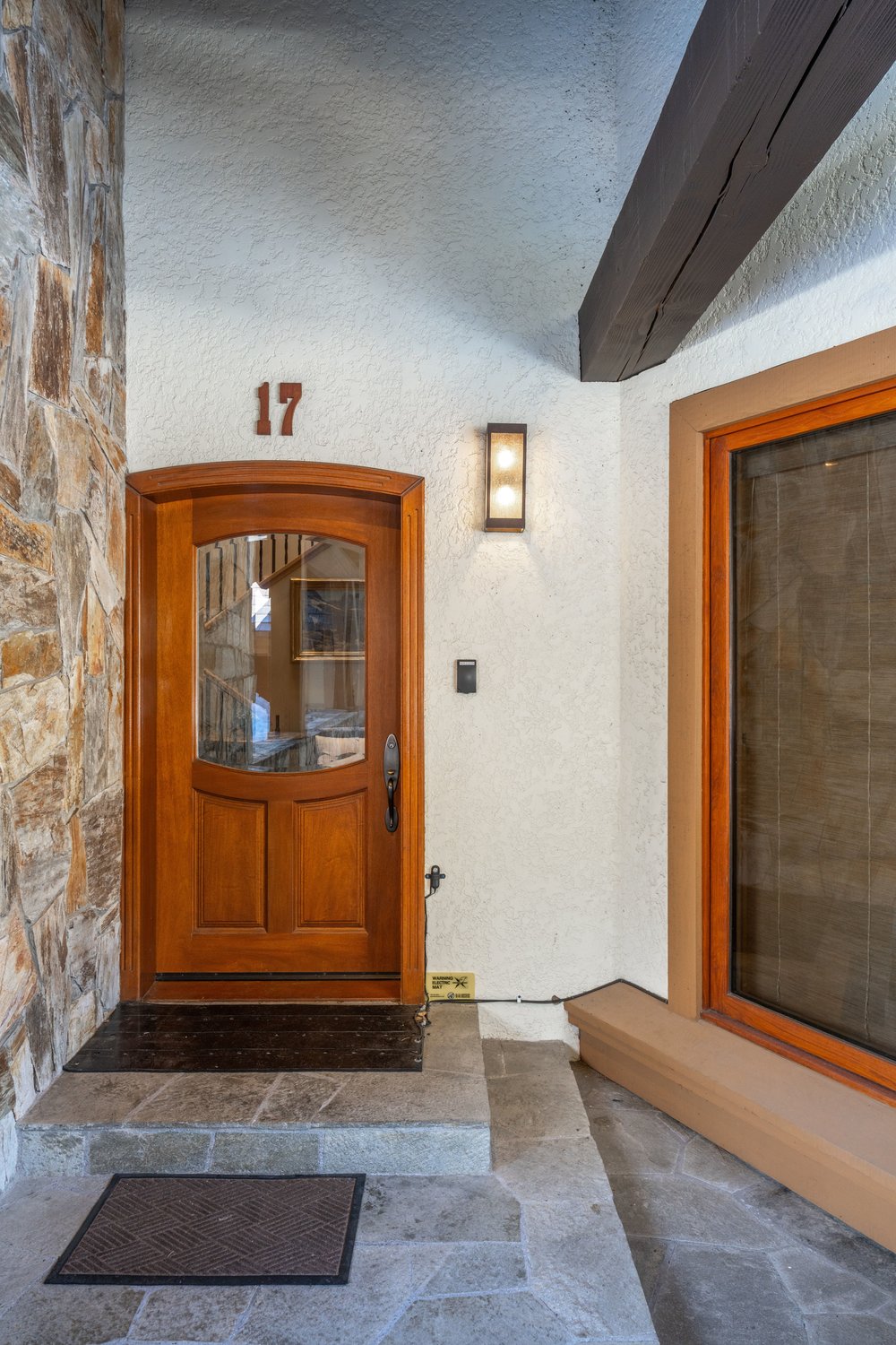 This image showcases the entryway of a home, featuring a wooden front door with glass panels and the number '17' above it. The exterior wall is a mix of stone and textured stucco, complemented by a modern light fixture. The entrance is elevated with stone steps and a doormat, creating a welcoming and stylish first impression.