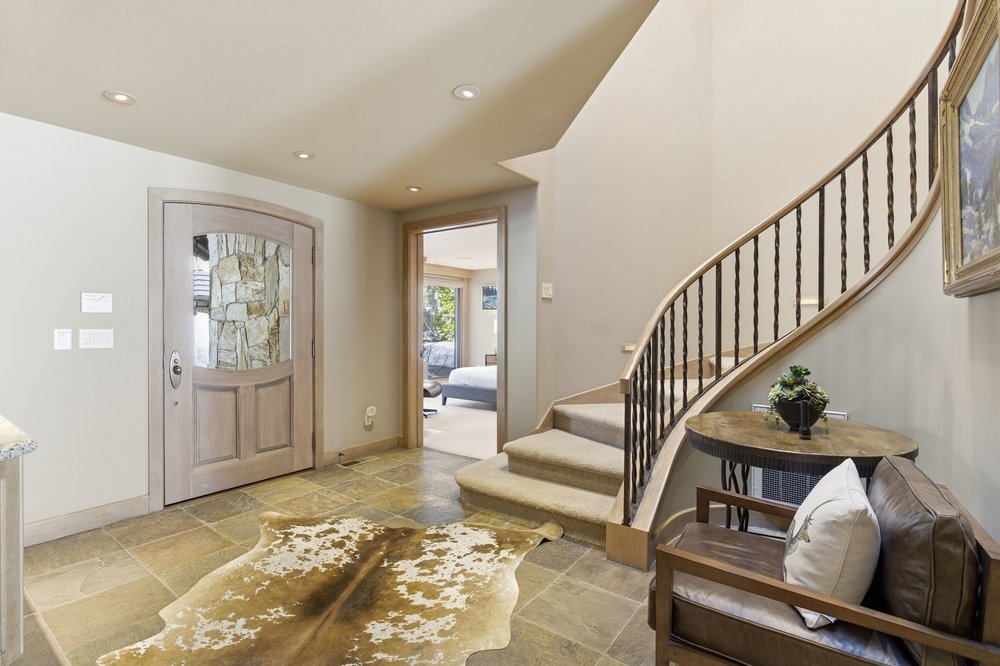 This interior shot showcases a welcoming hallway with a curved staircase. The flooring is tiled, and a decorative cowhide rug adds texture. A wooden door with a stone-accented window is visible, along with a glimpse into another room, suggesting a bedroom. The staircase features dark metal railings and leads upwards, creating a sense of flow and movement within the home.