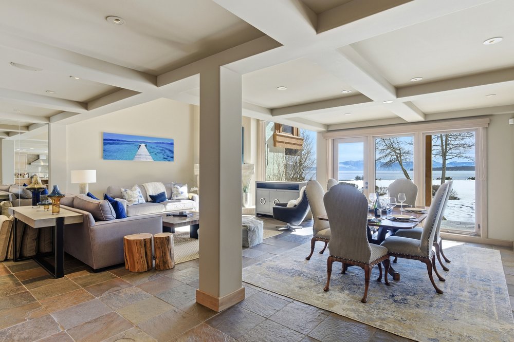 This interior shot showcases a combined living and dining area, featuring a comfortable gray sofa with blue accent pillows, a modern dining table set for six, and large windows offering a scenic view of a snowy landscape. The room is divided by a structural column, and the flooring is a mix of tile and area rug, creating a warm and inviting atmosphere perfect for relaxation and entertaining.