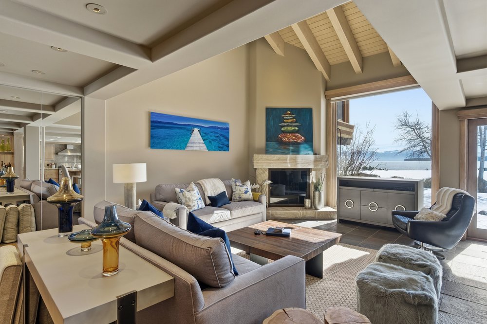 This is an interior shot of a living room featuring a neutral color palette, comfortable seating, and a fireplace. The room is well-lit with natural light from a large window offering a view of the water. Artwork adorns the walls, and the overall style is modern and inviting.