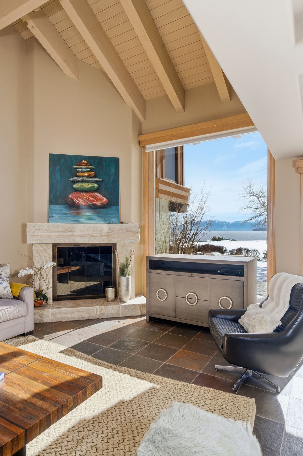 This is an interior shot of a living room featuring a stone fireplace with a painting above it, a modern cabinet, and a black leather chair. The room has a high, vaulted ceiling with exposed wooden beams and a large window offering a view of a snowy landscape. The overall style is modern and cozy, with a focus on natural light and comfortable furnishings.
