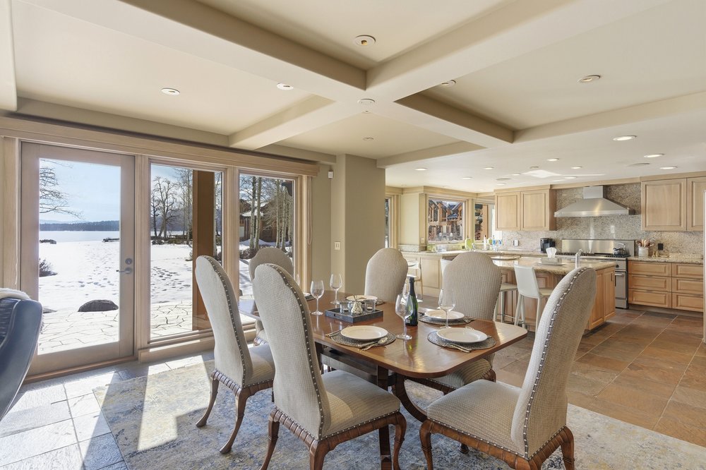 This interior shot showcases a dining room adjacent to a kitchen, featuring a wooden dining table set for a meal with upholstered chairs. Large glass doors offer a view of a snowy landscape, bringing natural light into the space. The room is decorated in neutral tones, creating a warm and inviting atmosphere.