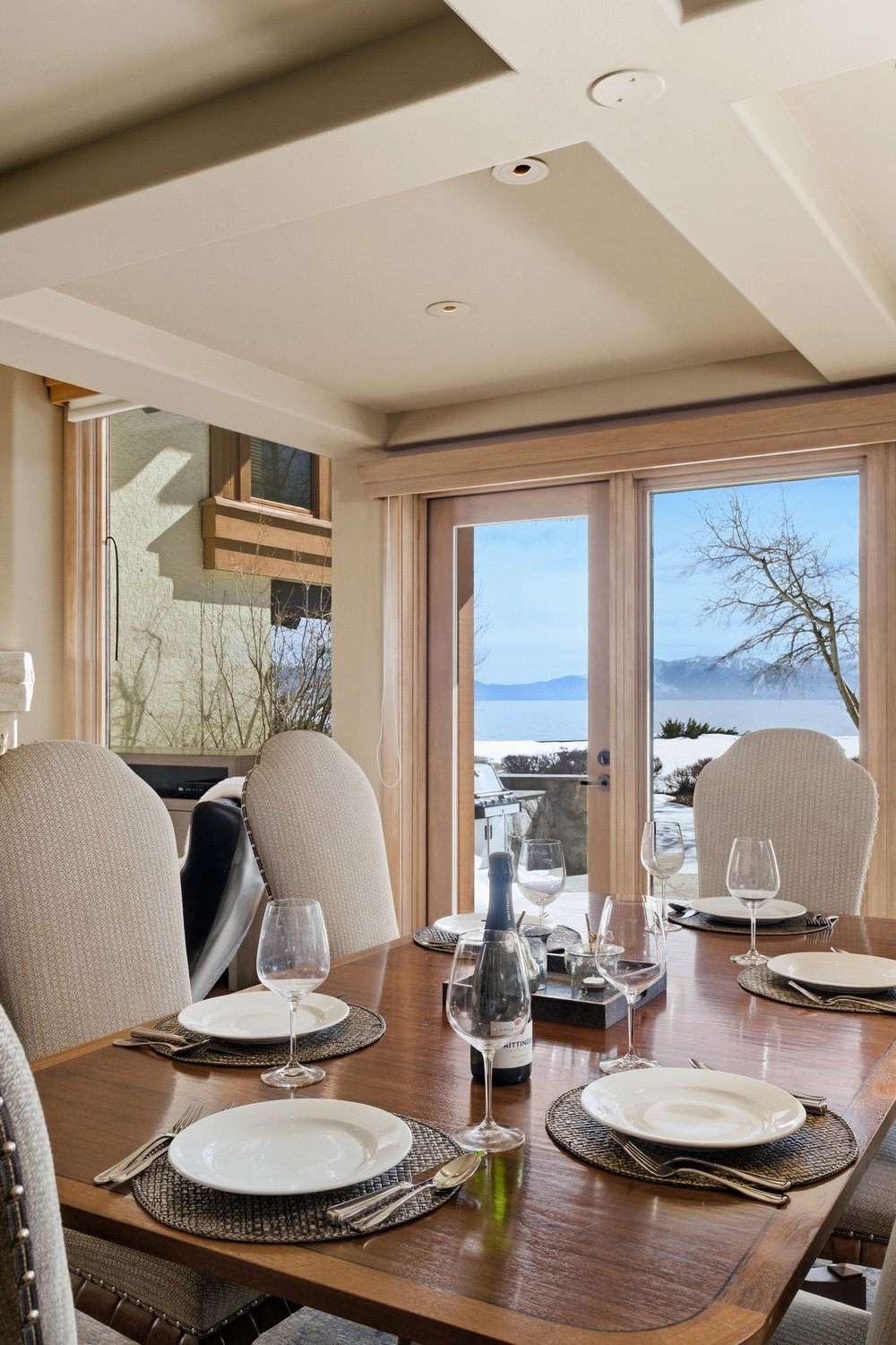 This is an interior shot of a dining room featuring a wooden dining table set for a meal. The table is adorned with plates, silverware, and wine glasses, suggesting a formal dining setting. Large windows provide a view of a snowy landscape, enhancing the room's ambiance and connection to the outdoors.