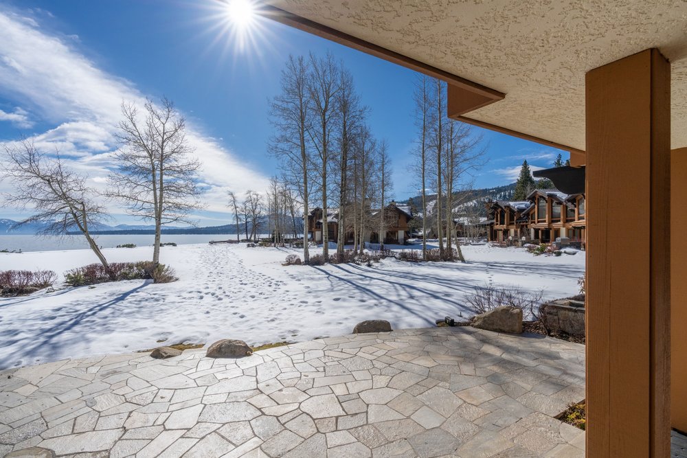 This image showcases a stone patio extending from a building, offering a picturesque view of a snow-covered landscape with bare trees and a lake in the distance. The patio's textured surface contrasts with the smooth snow, creating visual interest. The scene is bathed in sunlight, enhancing the serene and inviting atmosphere, perfect for outdoor relaxation and enjoyment of the natural surroundings.