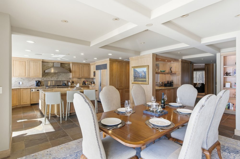 This interior shot showcases a dining room adjacent to a kitchen area. The dining room features a wooden table set for a meal, surrounded by upholstered chairs. The kitchen area includes wooden cabinetry, stainless steel appliances, and a breakfast bar, creating an open and inviting space.