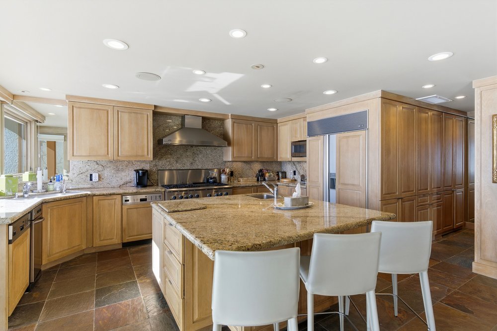 This is a well-lit kitchen featuring light wood cabinetry, granite countertops, and stainless steel appliances. A large island with seating dominates the center of the room, and the floor is tiled with earth-toned stone. The kitchen appears spacious and functional, with ample storage and counter space.