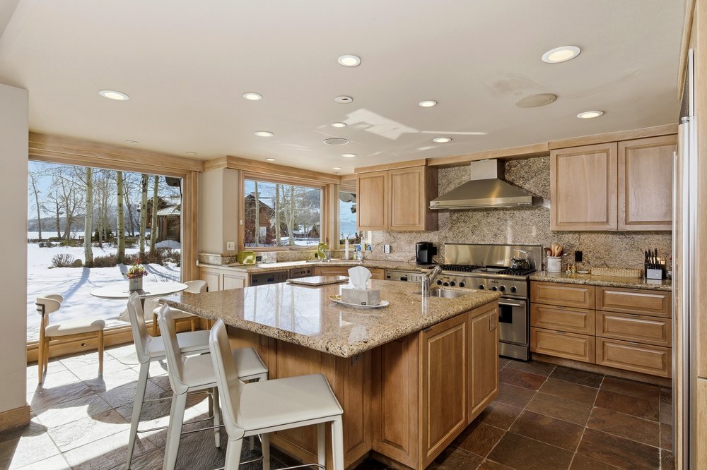 This is a well-lit kitchen featuring light wood cabinetry, granite countertops, and stainless steel appliances. A large island with bar seating anchors the space, while large windows offer a view of a snowy landscape. The kitchen has a warm and inviting feel, perfect for both cooking and entertaining.