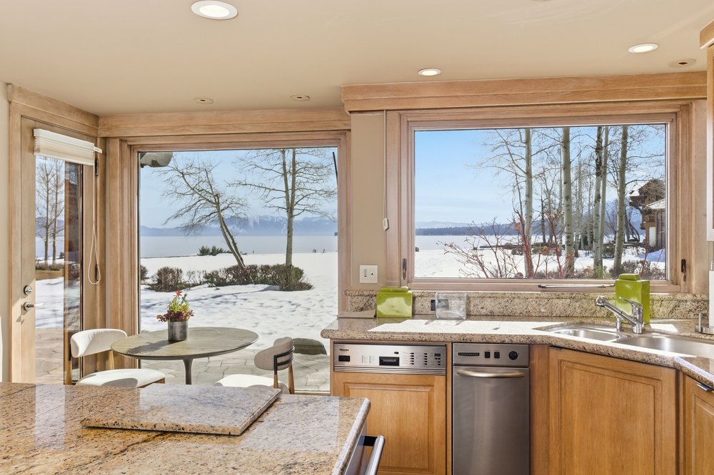 This kitchen features granite countertops, wooden cabinets, and stainless steel appliances, including a dishwasher. Large windows offer a scenic view of a snow-covered landscape with trees and a body of water. The kitchen is well-lit with recessed lighting, creating a warm and inviting atmosphere.