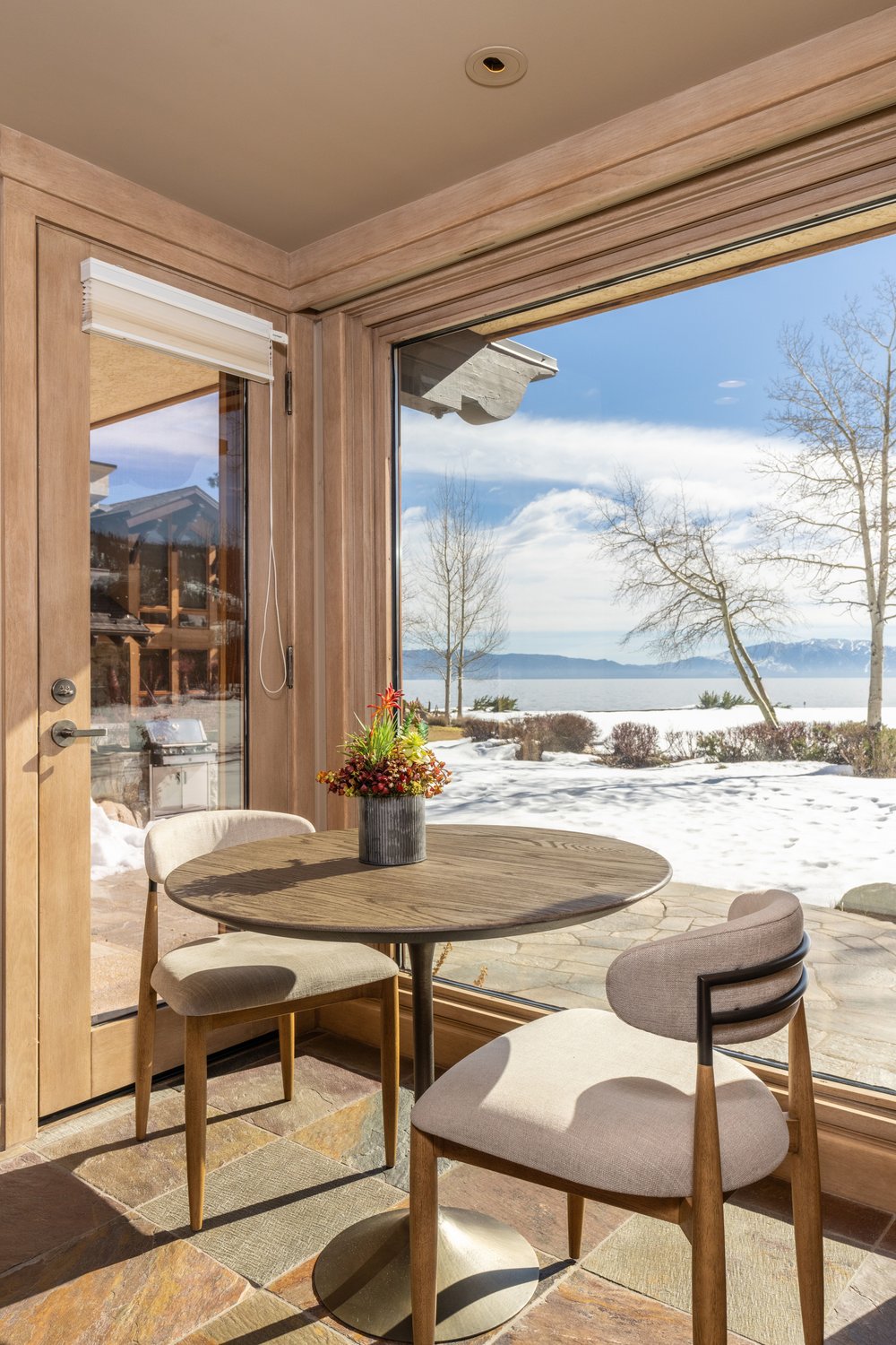 This interior shot showcases a cozy dining area with a round wooden table and two upholstered chairs. Large windows offer a scenic view of a snow-covered landscape and a body of water, creating a serene atmosphere. The room features natural wood trim and stone flooring, adding to the rustic yet elegant ambiance.