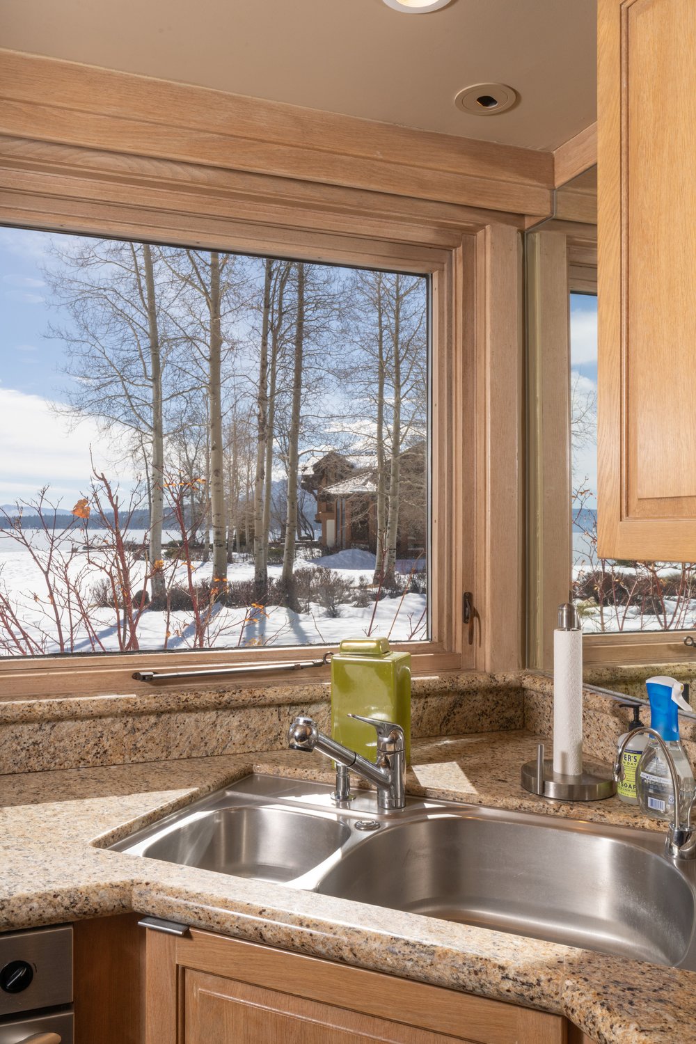 A kitchen sink area is showcased with a double stainless steel sink, a modern faucet, and a granite countertop. A window above the sink offers a view of a snow-covered landscape with bare trees and a glimpse of a house in the distance. The cabinetry is light wood, complementing the countertop.