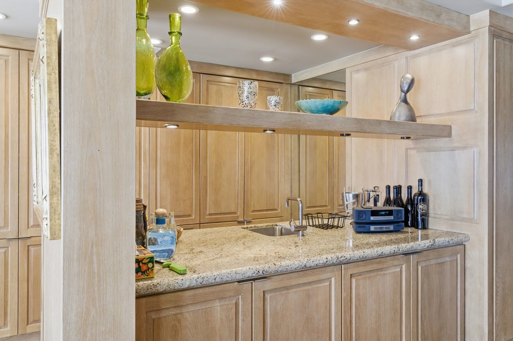 This image showcases a well-appointed kitchen area with light wood cabinetry and granite countertops. A wooden shelf displays decorative items, while the countertop features a sink, faucet, and various bottles and appliances. The overall impression is one of warmth and functionality, suggesting a comfortable and inviting space for cooking and entertaining.