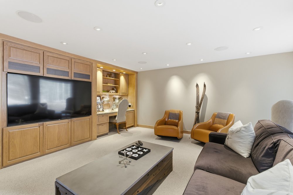 This interior shot showcases a cozy living room with built-in wooden cabinetry and a large television. The room features comfortable seating, including a brown sofa and two tan armchairs, arranged around a modern coffee table. The neutral color palette and recessed lighting create a warm and inviting atmosphere, perfect for relaxation and entertainment.