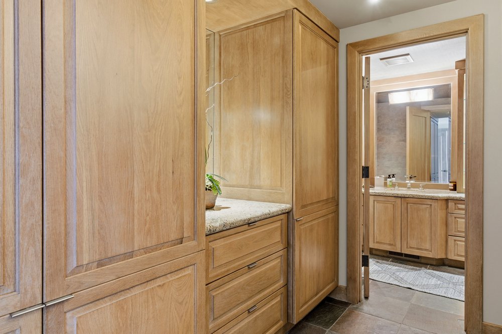 This is a well-lit bathroom featuring light wood cabinetry with granite countertops. The bathroom has a clean and modern design, with a view into another section of the bathroom through a doorway. The flooring appears to be tile, adding to the sophisticated aesthetic.