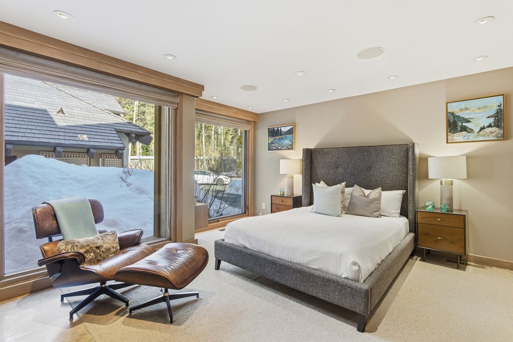 This is a primary bedroom featuring a large bed with a gray upholstered headboard and white bedding. The room has a neutral color palette with beige walls and carpet. Large windows offer natural light and views of the snowy exterior, complemented by a comfortable leather chair and ottoman. Artwork adorns the walls, and bedside tables with lamps provide soft lighting.