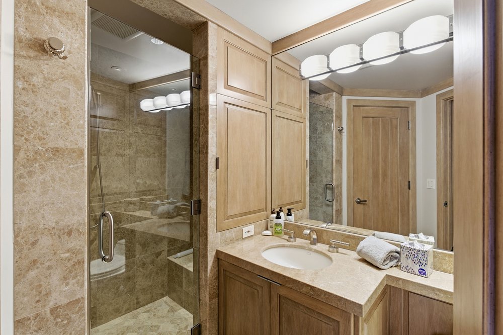 This is a primary bathroom featuring a glass-enclosed shower with marble-like tile, a vanity with light wood cabinetry and a stone countertop, and a large mirror with overhead lighting. The bathroom has a clean and luxurious feel, with a neutral color palette and modern fixtures. The shower is spacious and well-lit, and the vanity provides ample storage space.
