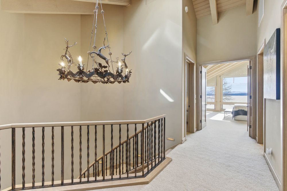This interior shot showcases a hallway with stairs, featuring a unique chandelier with deer figures. The staircase has a wooden railing with dark metal spindles, and the hallway leads to a bright room with a view of a lake. The walls are painted in a neutral tone, and the carpet is light-colored, creating a warm and inviting atmosphere.