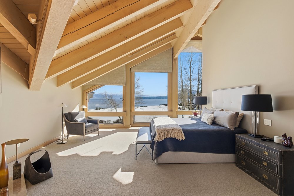 This is a primary bedroom featuring exposed wooden beams and large windows with a view of a lake. The room is furnished with a bed, nightstand, and armchair, creating a cozy and inviting atmosphere. The neutral color palette and natural light enhance the serene ambiance, making it a desirable space for relaxation.