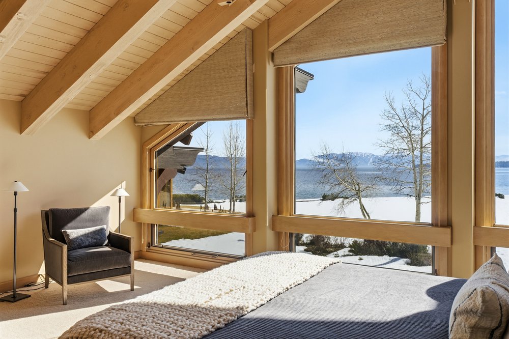 This primary bedroom features large windows with views of a snow-covered landscape and a lake. The room has a vaulted ceiling with exposed wooden beams, a gray upholstered armchair, and a bed with a gray coverlet and a chunky knit throw. The overall style is cozy and luxurious, emphasizing the natural light and scenic views.