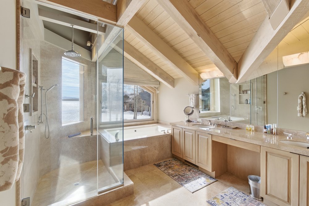 This is a luxurious primary bathroom featuring a glass-enclosed shower with a rainfall showerhead and a separate soaking tub. The vanity area includes dual sinks with light wood cabinetry and a light-colored countertop. The room has a bright and airy feel, enhanced by natural light streaming through the windows, and the exposed wooden beams add a touch of rustic charm.