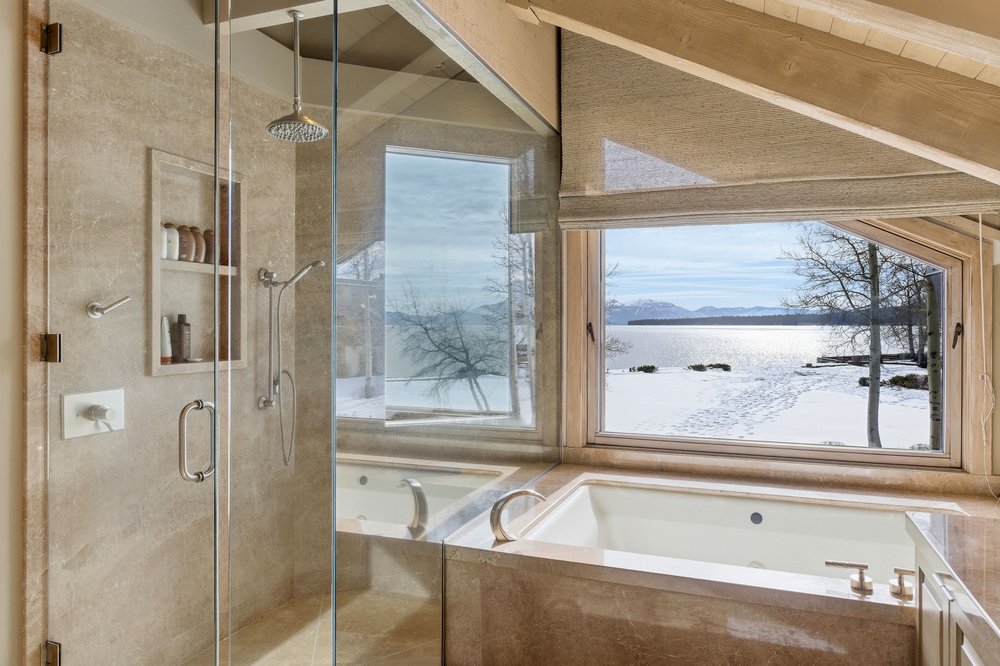 This is a luxurious primary bathroom featuring a glass-enclosed shower and a soaking tub with a stunning view of a snow-covered landscape and lake. The bathroom is finished with neutral-toned stone or tile, creating a spa-like atmosphere. The large window provides ample natural light and highlights the scenic backdrop, making this a relaxing and visually appealing space.