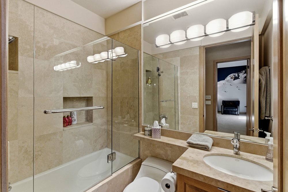 This is a well-lit bathroom featuring beige marble tiling throughout. A glass-enclosed bathtub and shower combination is visible, along with a toilet and a vanity with a marble countertop and wooden cabinets. The large mirror reflects the bathroom and a glimpse into an adjacent room, creating a sense of spaciousness.