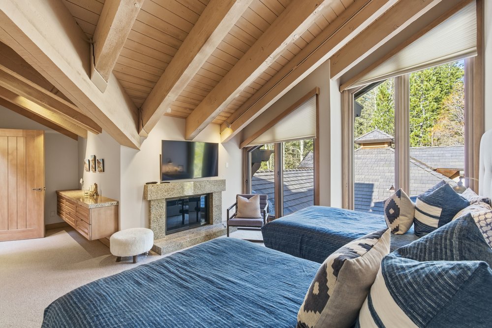 This is a cozy guest bedroom featuring two twin beds with blue bedding and decorative pillows. The room has a vaulted ceiling with exposed wooden beams, a stone fireplace with a mounted TV, and large windows offering natural light and views of the outdoors. A small desk area and a comfortable chair add to the room's functionality and charm.