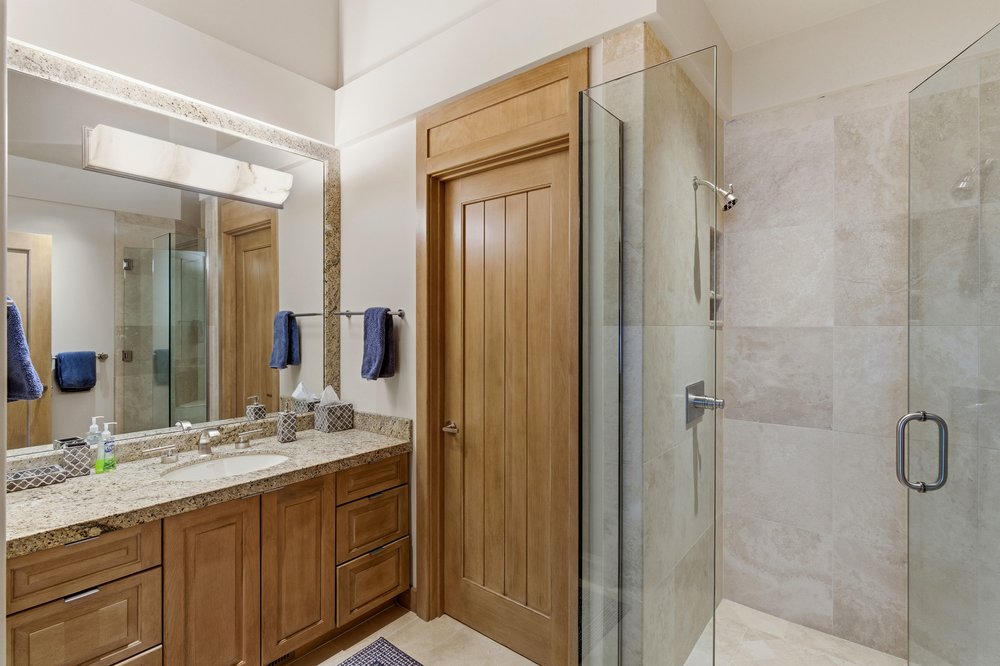 This is a well-lit primary bathroom featuring a wooden vanity with a granite countertop and a large mirror with integrated lighting. A glass-enclosed shower with neutral-toned tiles is adjacent to the vanity. The overall impression is clean, modern, and luxurious.