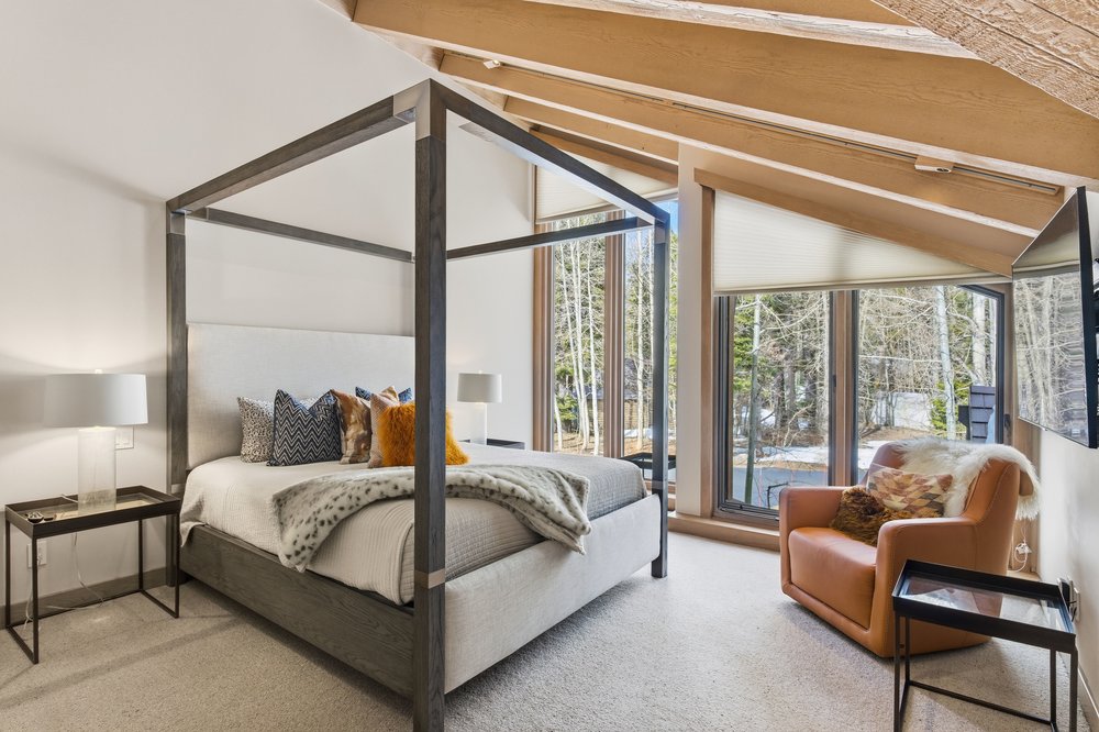 This is a primary bedroom featuring a four-poster bed with neutral-toned bedding and decorative pillows. The room has large windows offering natural light and views of the surrounding trees. An armchair and side tables add to the comfort and functionality of the space, creating a serene and inviting atmosphere.