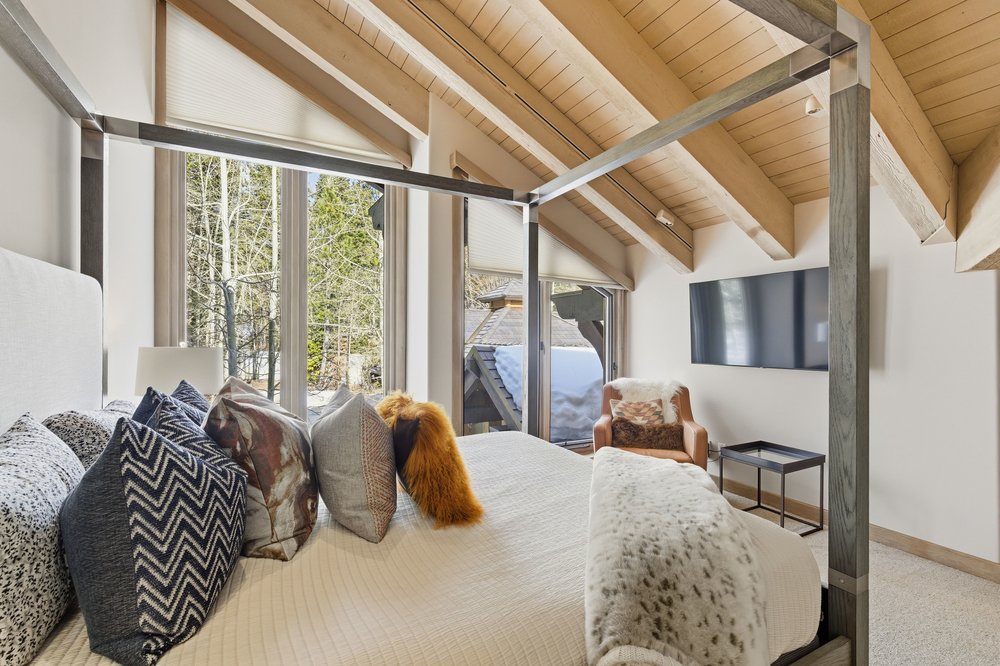 This is a primary bedroom featuring a four-poster bed with a light-colored comforter and decorative pillows. The room has a vaulted ceiling with exposed wooden beams and large windows that provide natural light. A television is mounted on the wall, and a comfortable chair sits in the corner, creating a cozy and inviting atmosphere.