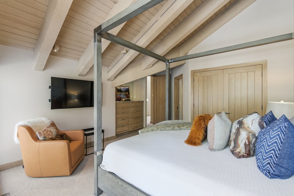 This is a primary bedroom featuring a four-poster bed with white linens and decorative pillows. A tan armchair sits to the side of the bed, facing a wall-mounted television. The room has a vaulted ceiling with exposed beams, adding to the cozy and luxurious feel.