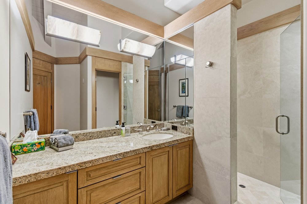 This is a well-lit primary bathroom featuring a granite countertop vanity with wooden cabinets and a circular sink. A large mirror reflects the vanity area and a nearby doorway. A glass-enclosed shower is visible to the right, showcasing a modern and clean design.
