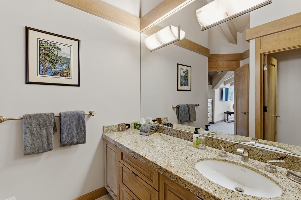 This is a well-lit bathroom featuring a granite countertop vanity with wooden cabinets and drawers. A large mirror reflects the vanity and the opposite wall, which is adorned with a framed picture and a towel rack holding two gray towels. The bathroom has a neutral color palette with wooden beams on the ceiling, creating a warm and inviting atmosphere.