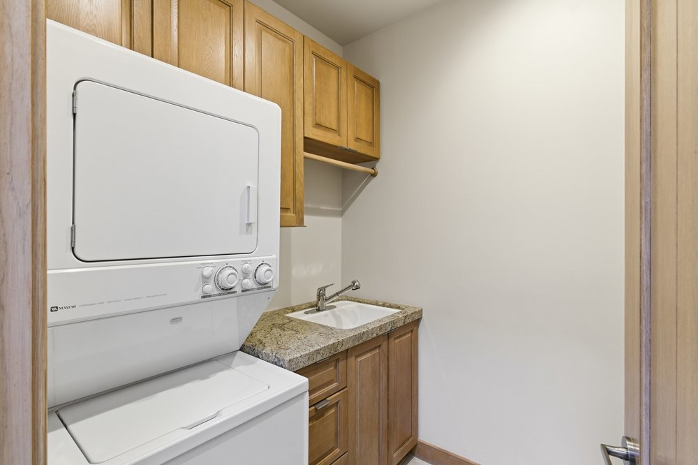 This is an interior shot of a laundry room. It features a stacked washer and dryer unit, wooden cabinets above and below a granite countertop with a sink. The room is well-lit and appears functional, ideal for a real estate listing highlighting home amenities.