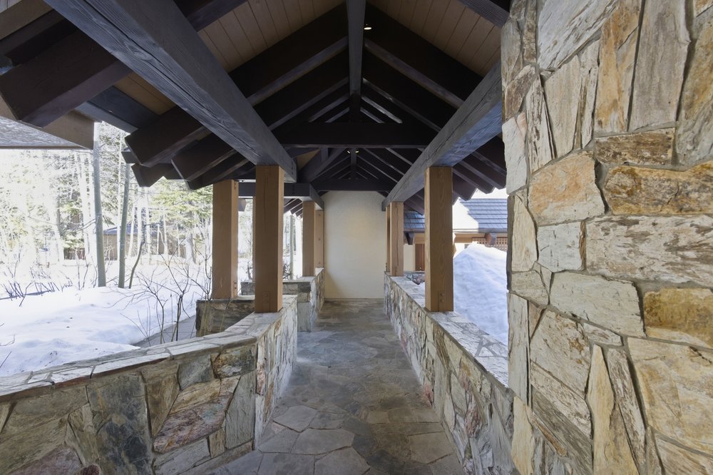 This image showcases a covered entryway with a rustic and inviting design. The structure features exposed dark wood beams and supports, complemented by natural stone walls and flooring. The entryway provides shelter from the elements and leads to the main entrance of the property, with a snowy landscape visible in the background.